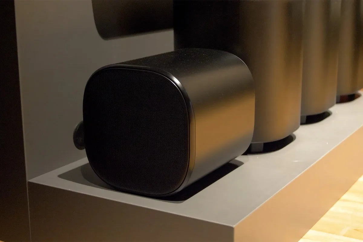 Een zwarte subwoofer van de JBL Bar 1300MKII staat prominent op een grijze standaard. Het ontwerp is afgerond en minimalistisch, met een matzwarte afwerking.
