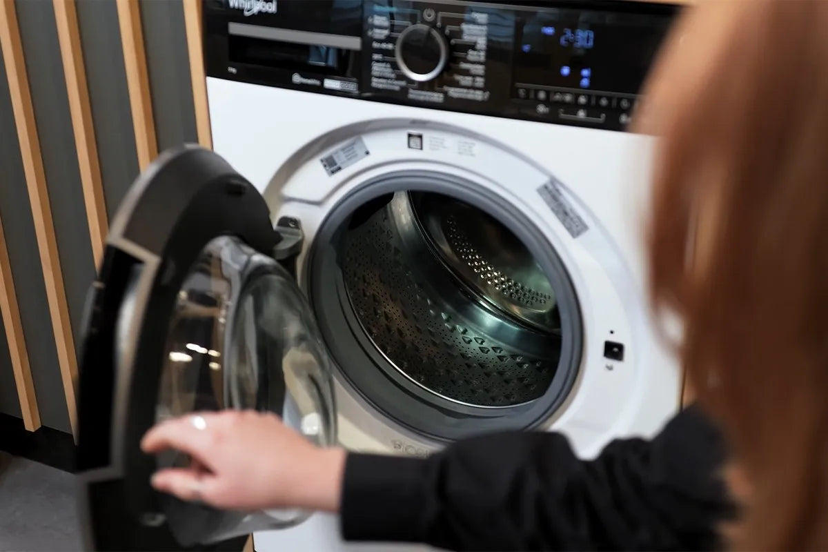 Een vrouw opent de deur van een Whirlpool-wasmachine; het display toont een resterende wastijd van 2:30 uur en de binnenkant van de trommel is goed zichtbaar met een karakteristiek patroon op het metaal.