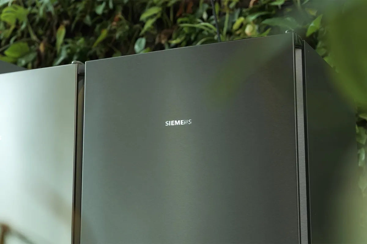 Tussen de bladeren van een plant door is de voorkant van een Siemens iQ300-koelkast te zien.