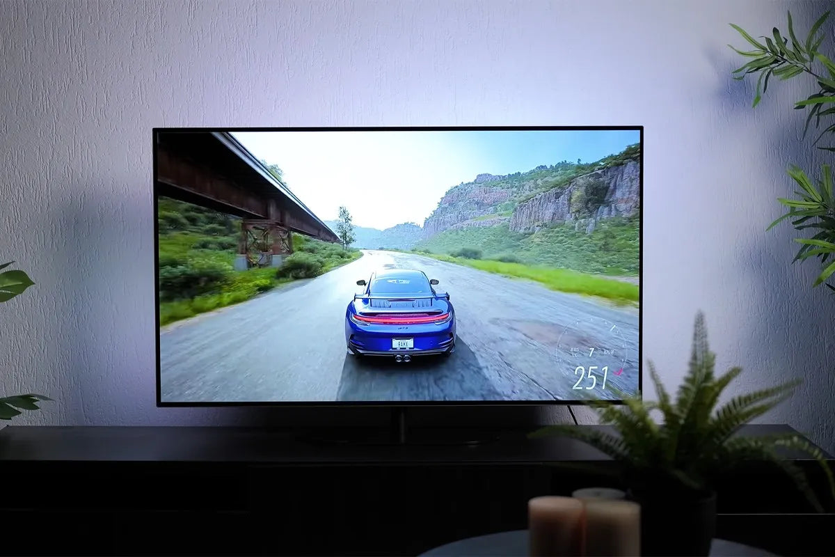 Philips OLED-tv waarop een racespel wordt gespeeld met een blauwe sportwagen die met hoge snelheid (251 km/u) over een weg rijdt langs rotswanden en een spoorbrug; het camerastandpunt is achter de auto, en de muur achter de tv is subtiel blauw verlicht door Ambilight-verlichting.
