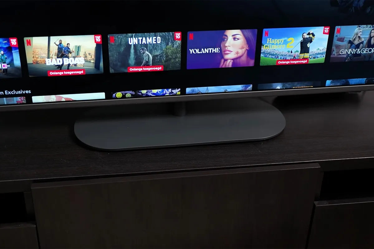 Een televisie die op een donker tv-meubel staat, met op het scherm het Netflix-menu waarop diverse titels worden getoond zoals “BAD BOAS”, “UNTAMED”, “YOLANTHE”, “Happy Gilmore 2” en “GINNY & GEORGIA”, en met in het midden van het beeld de ovale voet van de tv goed zichtbaar