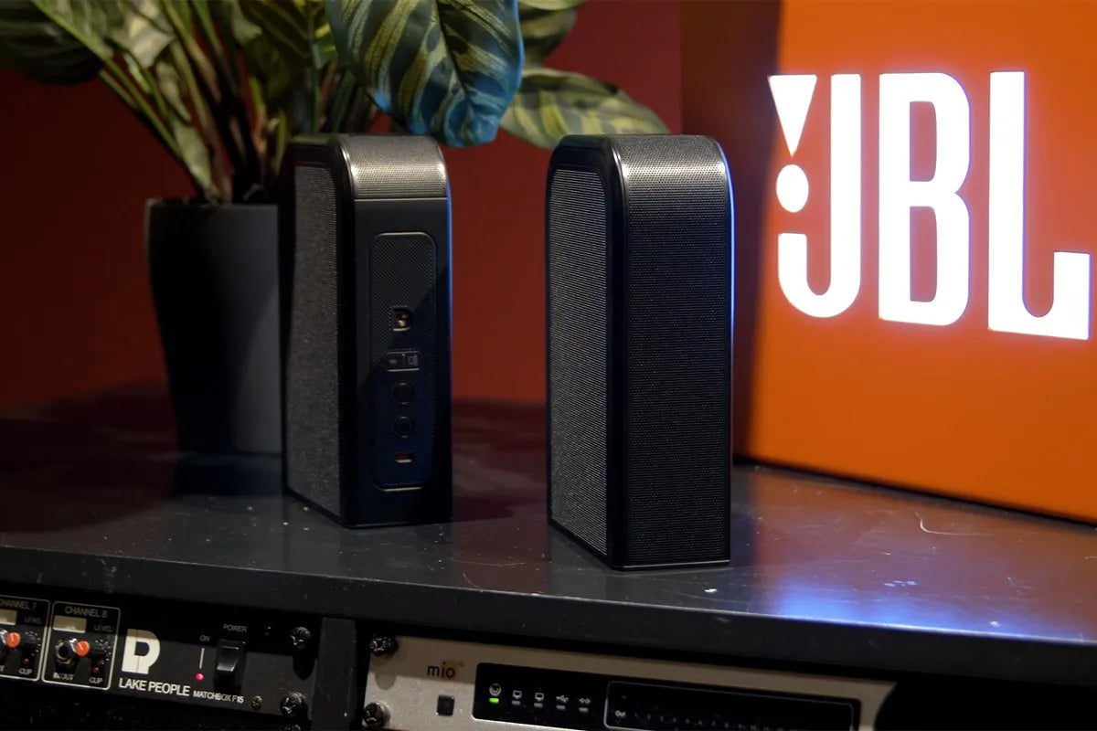 Twee losse speakers van de JBL Bar 1300MKII staan rechtop op een meubel, naast een oplichtend JBL-logo tegen een oranje achtergrond.