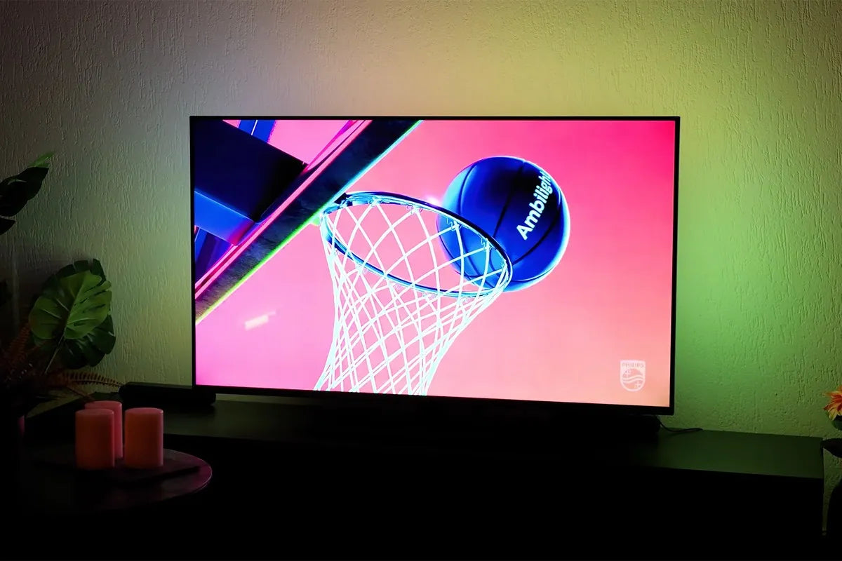 Philips The One 2025 waarop een kleurrijk beeld wordt weergegeven van een basketbal met het woord “Ambilight” die net door een basketbalring gaat, tegen een felroze achtergrond, terwijl de muur achter de tv verlicht wordt met groenige Ambilight-verlichting