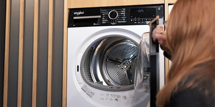 Een vrouw opent de deur van een Whirlpool-droger die is ingebouwd in een houten kastwand. De binnenkant van de metalen trommel is goed zichtbaar, inclusief het karakteristieke patroon van luchtopeningen en ribbels.