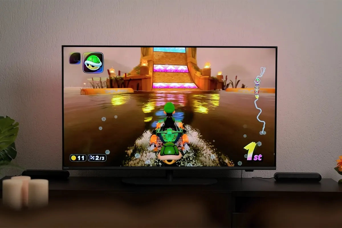 Philips The One 2025 waarop Mario Kart wordt gespeeld met personage Luigi dat op een wateroppervlak racet richting een tempelachtige helling met gekleurde lichtstroken; links onderin is te zien dat de speler 11 munten heeft en zich in ronde 2 van 3 bevindt, en rechts onderin staat dat hij op de eerste plaats ligt (“1st”), terwijl de muur subtiel verlicht wordt met Ambilight-verlichting.