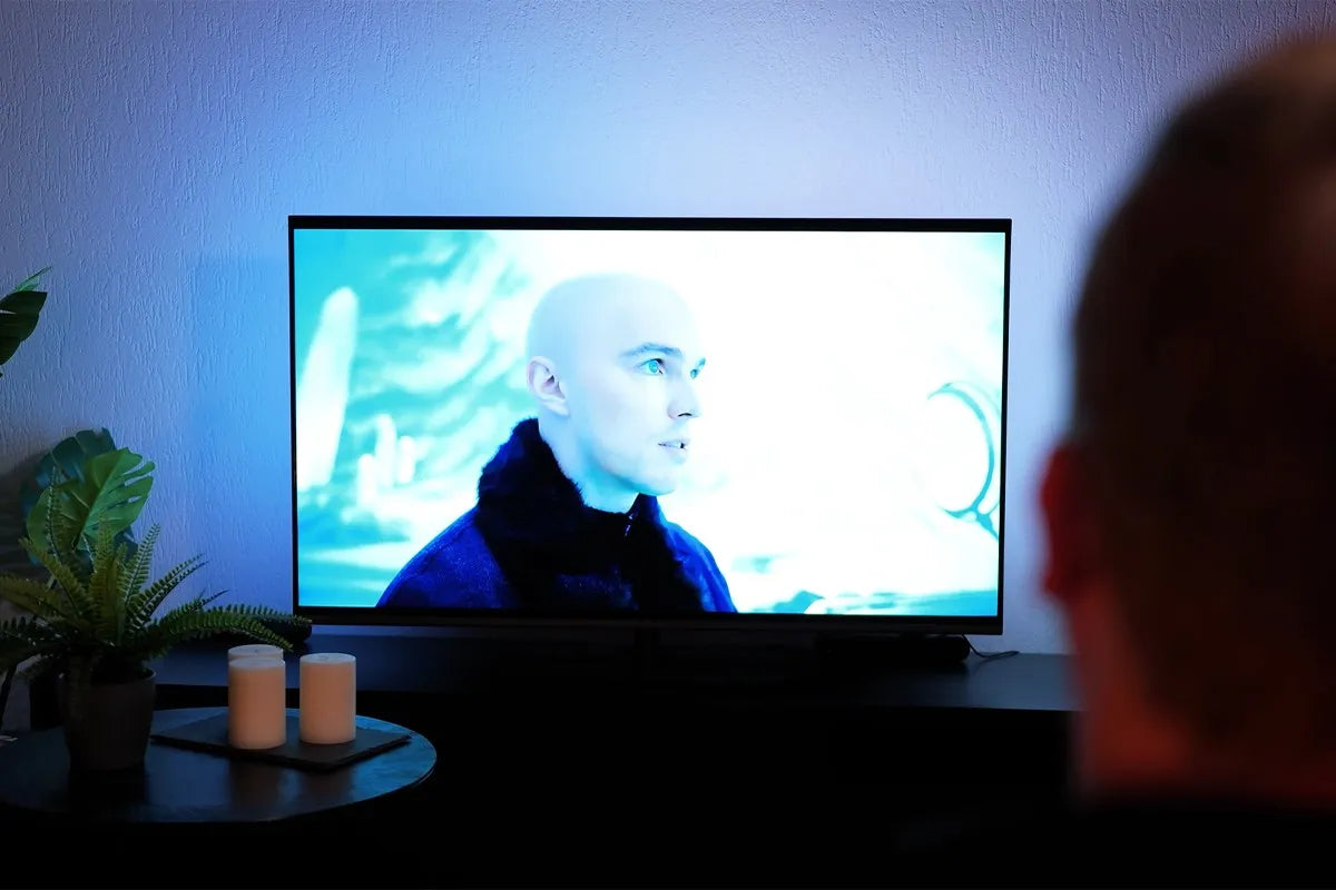 Philips The One 2025 waarop een scène wordt afgespeeld van een kale man in een blauw getinte omgeving uit een film, terwijl een persoon op de voorgrond met zijn rug naar de camera toe zit te kijken; de muur achter de tv wordt blauw verlicht door Ambilight