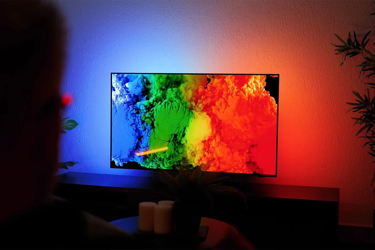 Philips OLED-tv toont een kleurrijke explosie van inktachtige wolken in blauw, groen, geel en rood op een zwarte achtergrond terwijl Ambilight-verlichting aan de muur.