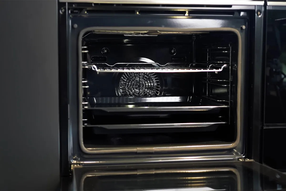 Een open inbouwoven met een schone, glanzende binnenkant; zichtbaar zijn een metalen rooster in het midden en een bakplaat eronder, met achterin de oven een ventilator voor heteluchtfunctie.