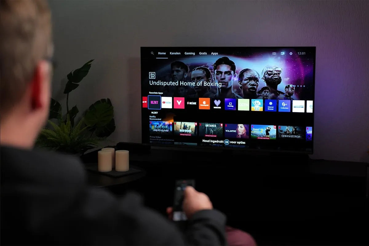 Een persoon zit op de bank en bedient met een afstandsbediening de Philips The One die het startscherm van Titan OS toont, met bovenaan het menu ‘Home’, ‘Kanalen’, ‘Gaming’, ‘Gratis’ en ‘Apps’, verder zijn apps zoals YouTube, NLZIET, Videoland en Netflix zichtbaar, en op de achtergrond is paarse Ambilight-verlichting te zien tegen de muur