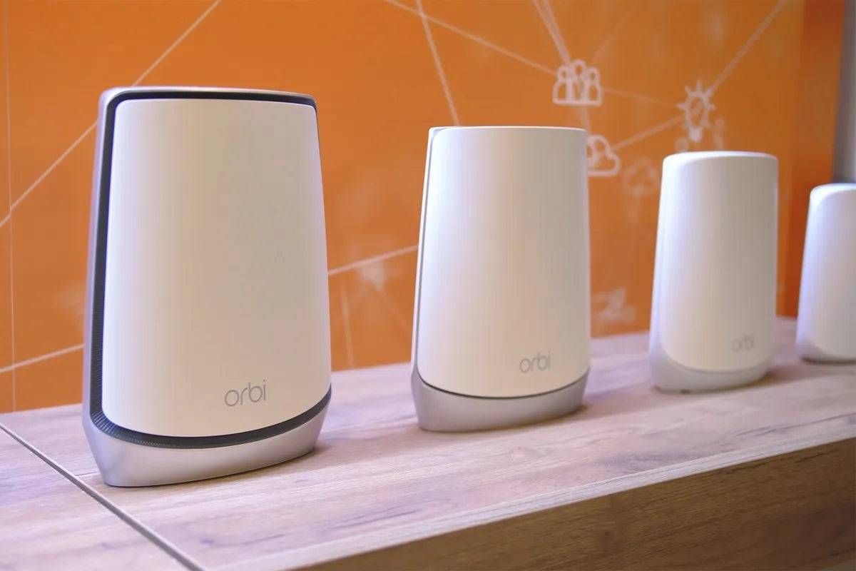 Vier Netgear Orbi wifi-routers staan opgesteld op een houten tafel met een oranje achtergrond.