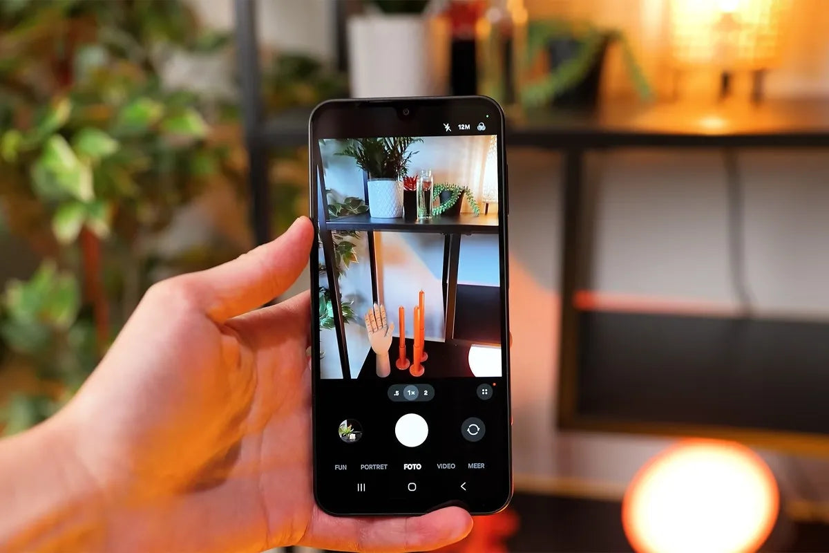 Een hand houdt een Samsung Galaxy A17-smartphone vast met de camera-app geopend in de fotomodus; op het scherm wordt een kleurrijk interieur vastgelegd met planten, rode kaarsen en decoratieve objecten op een metalen rek