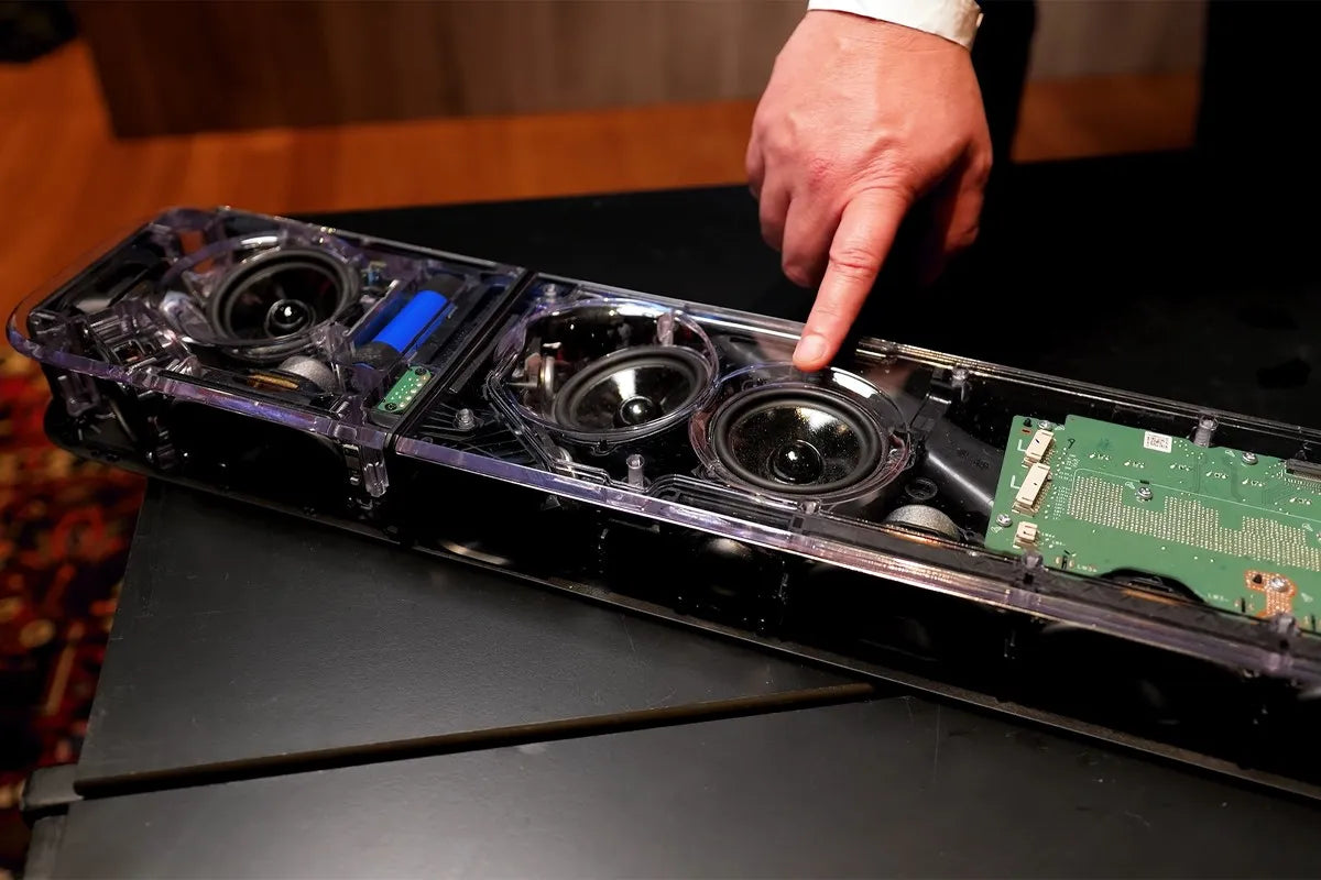 Een opengewerkte JBL Bar 1300MKII waarin de luidsprekers zichtbaar zijn. Een hand wijst naar een van de drivers, met een duidelijke focus op de interne techniek en hoogwaardige bouwkwaliteit.