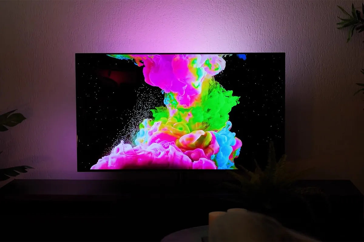Philips OLED-tv die een explosie van kleurrijke vloeistoffen toont in felle tinten roze, groen, geel en blauw op een zwarte achtergrond terwijl de muur achter de tv wordt verlicht met paarse Ambilight-verlichting die de levendige kleuren op het scherm versterkt.