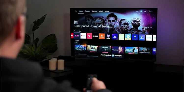Een persoon zit op de bank en bedient met een afstandsbediening de Philips The One die het startscherm van Titan OS toont, met bovenaan het menu ‘Home’, ‘Kanalen’, ‘Gaming’, ‘Gratis’ en ‘Apps’, verder zijn apps zoals YouTube, NLZIET, Videoland en Netflix zichtbaar, en op de achtergrond is paarse Ambilight-verlichting te zien tegen de muur