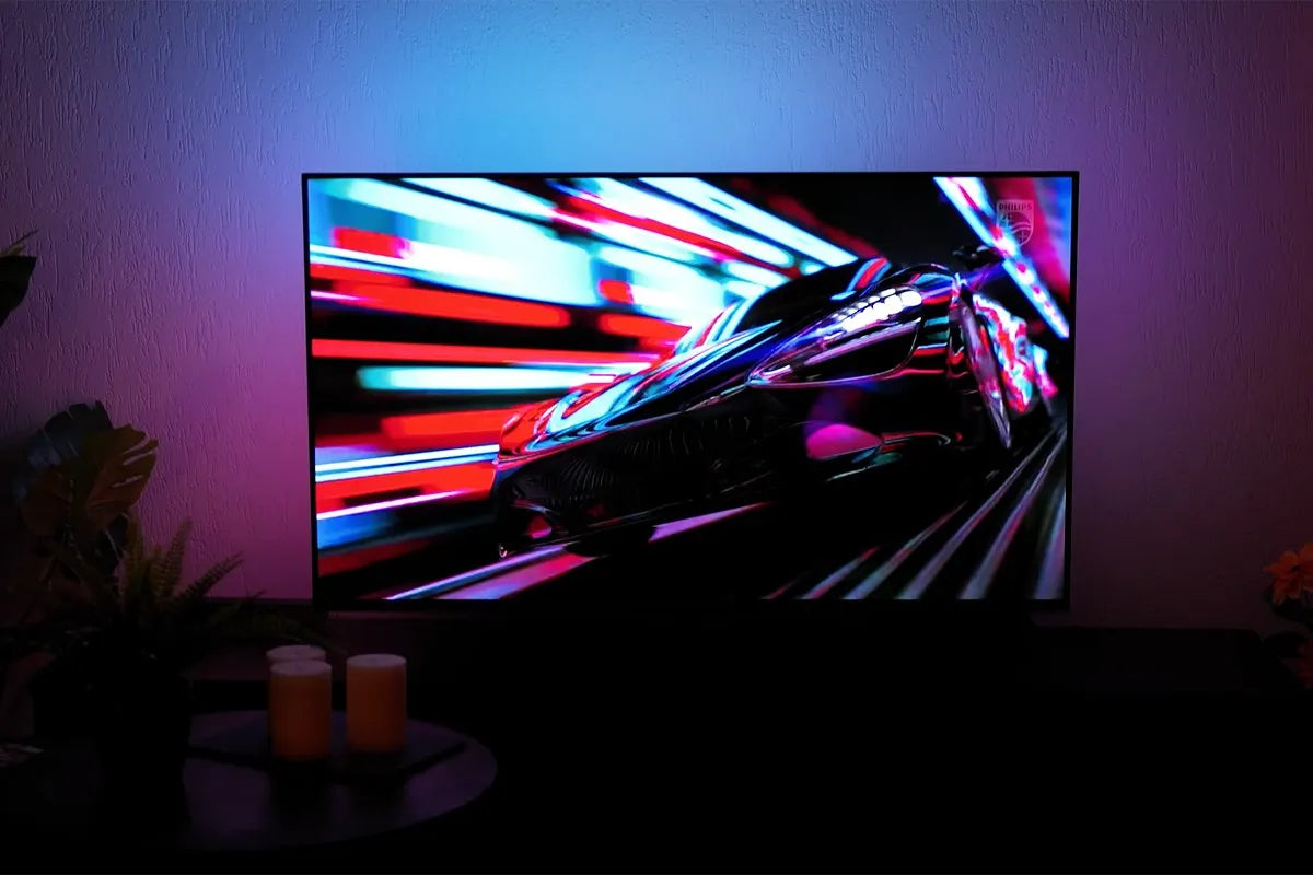 Philips The One 2025 waarop een futuristisch ogende sportwagen met hoge snelheid door een tunnel van kleurrijke lichtstroken raast, met felle rode, blauwe en paarse tinten die via Ambilight-verlichting sfeervol op de muur achter de tv worden geprojecteerd